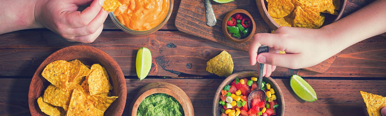 On a wooden table sits bowls of queso, salsa, guacamole and corn chips with hands reaching in to grab a serving On a wooden table sits bowls of queso, salsa, guacamole and corn chips with hands reaching in to grab a serving