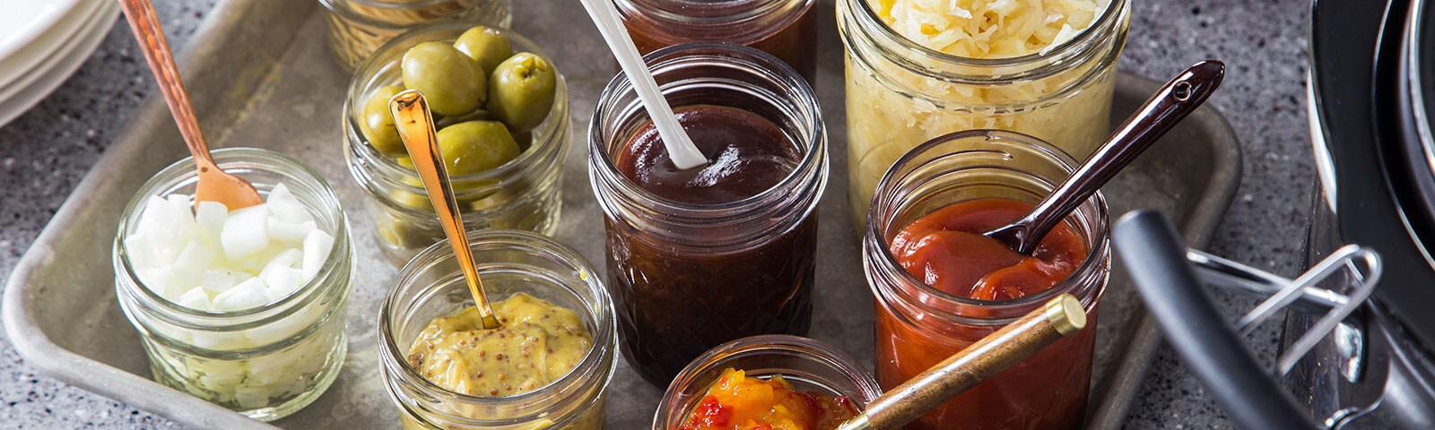 A bunch of condiments like ketchup, olives, BBQ sauce and sauerkraut on a metal tray