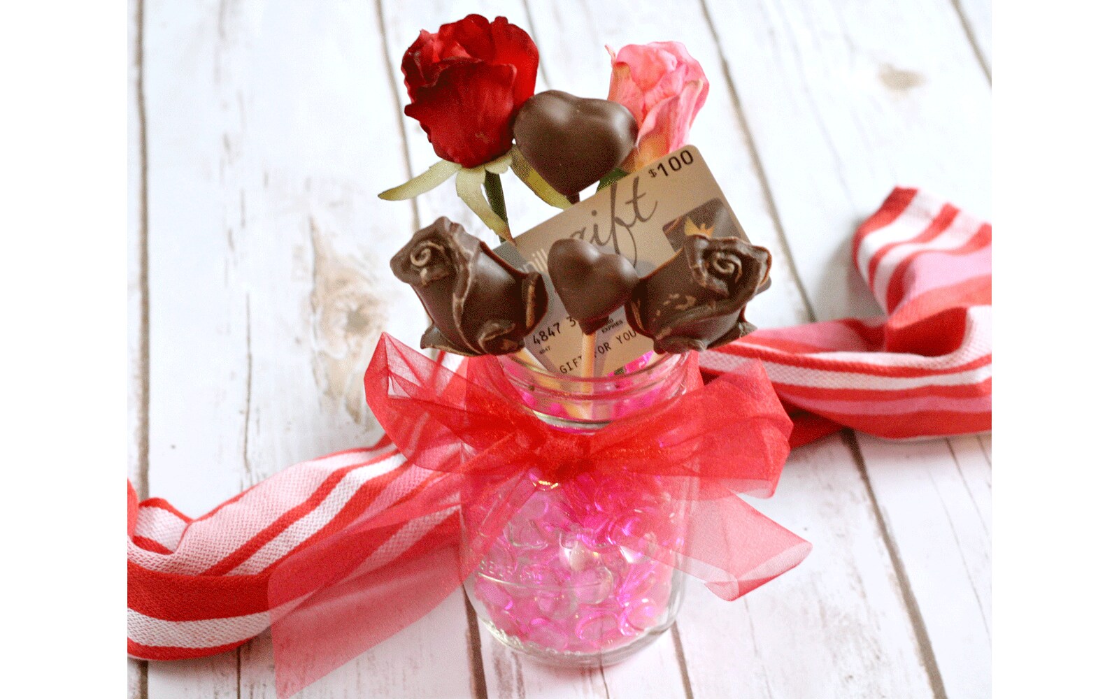 Mason jar wrapped with red ribbon tied into a bow and filled with chocolate on lollipop sticks