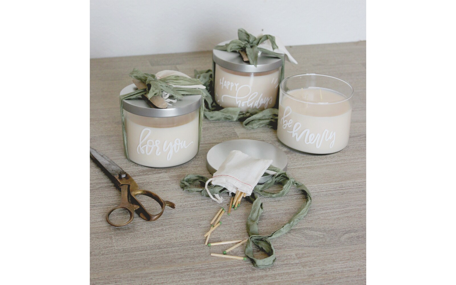 White candles with silver lids and words written on the glass  sit on a table next to scissors and matches