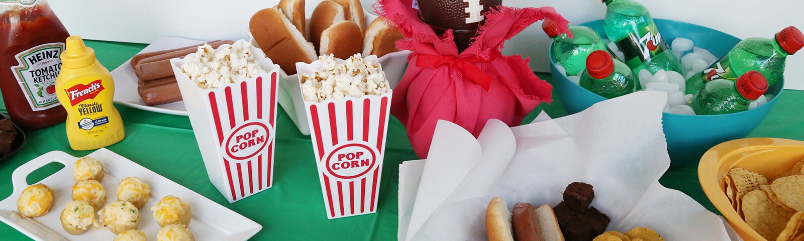 An array of football ready treats including popcorn, hotdogs and chips on a table covered with a green tablecloth